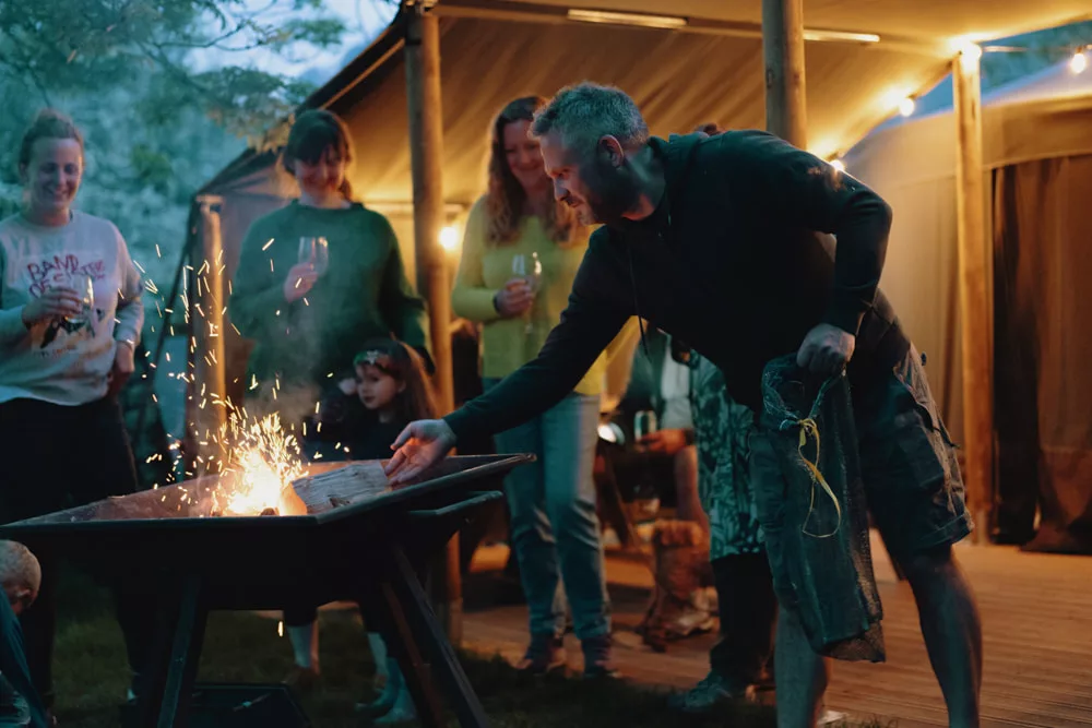 A man puts a log onto a fire pit, surrounded by a group of friends